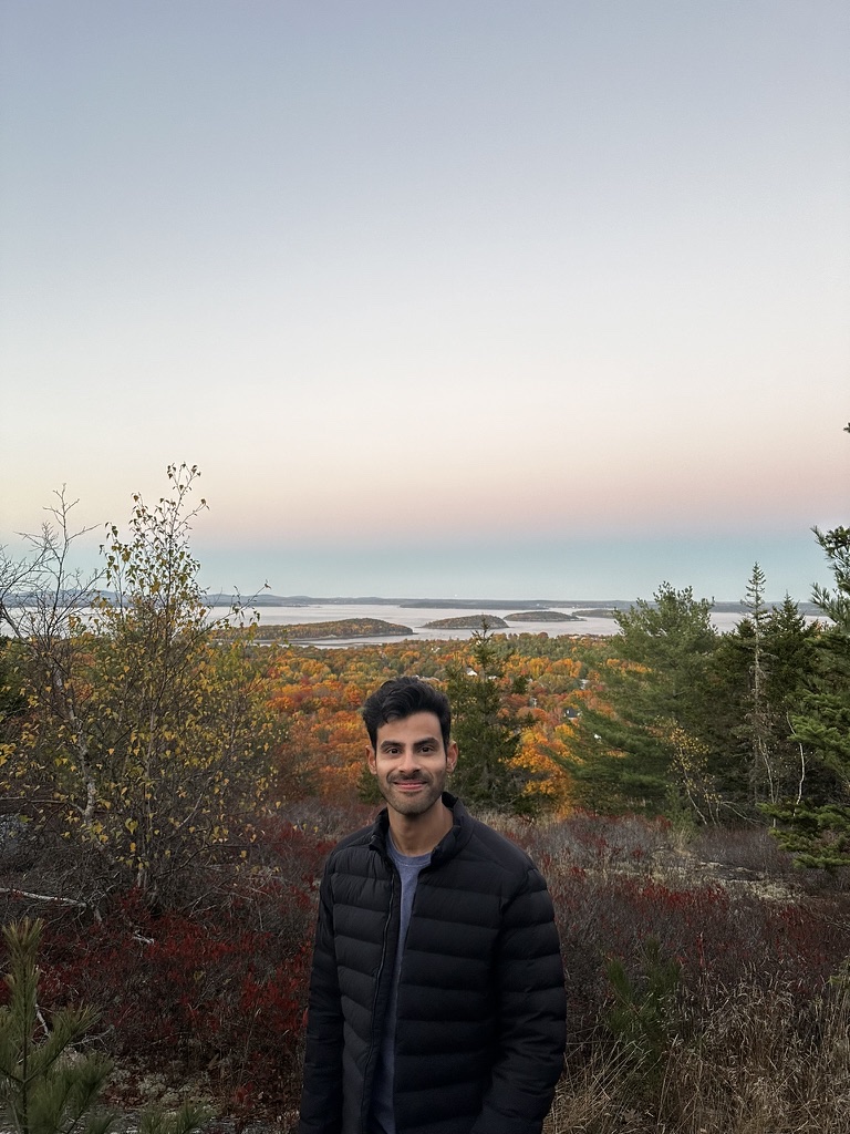 Ribhav Hora standing on a hillside in Acadia at dusk, fall foliage and islands behind him